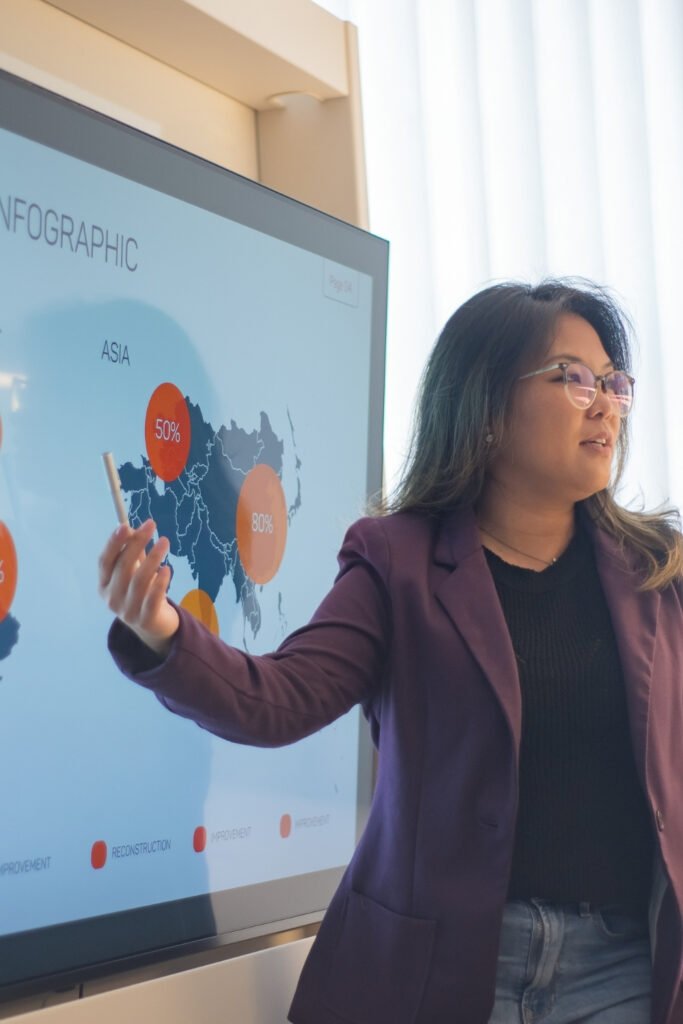 Non-designers need to turn words into visuals - asian marketing professional (woman) pointing at a large screen PowerPoint presentation.