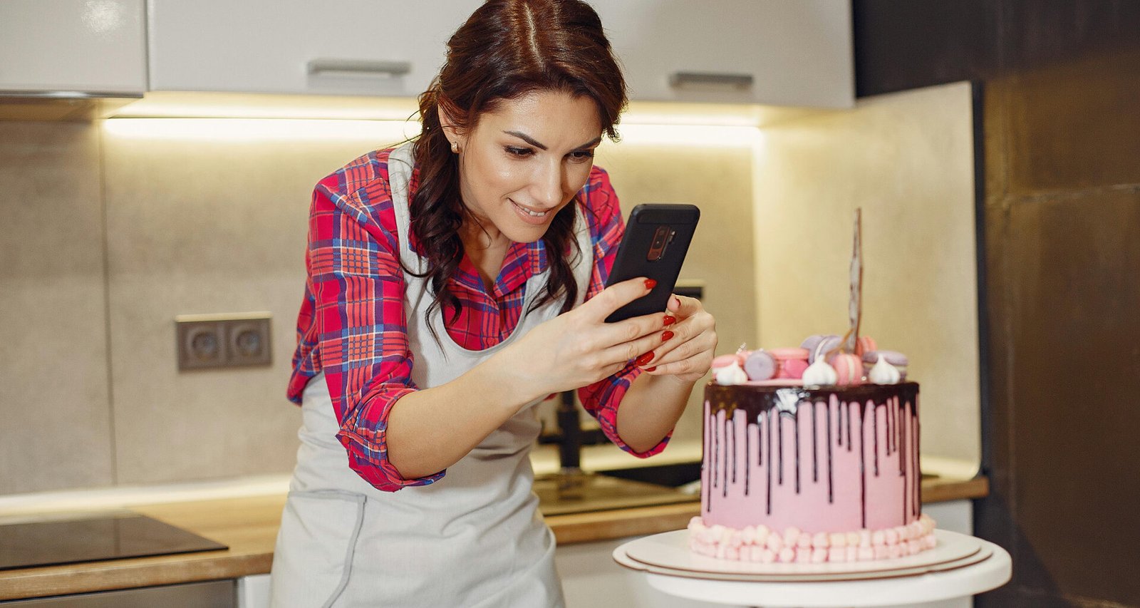 Feature image for the article: 'How to Know If Your Blog Idea Is Worth It (Before You Start)'. A young, white female, home baker, taking a photo of her decorated cake withe smart phone, kitchen in the background.